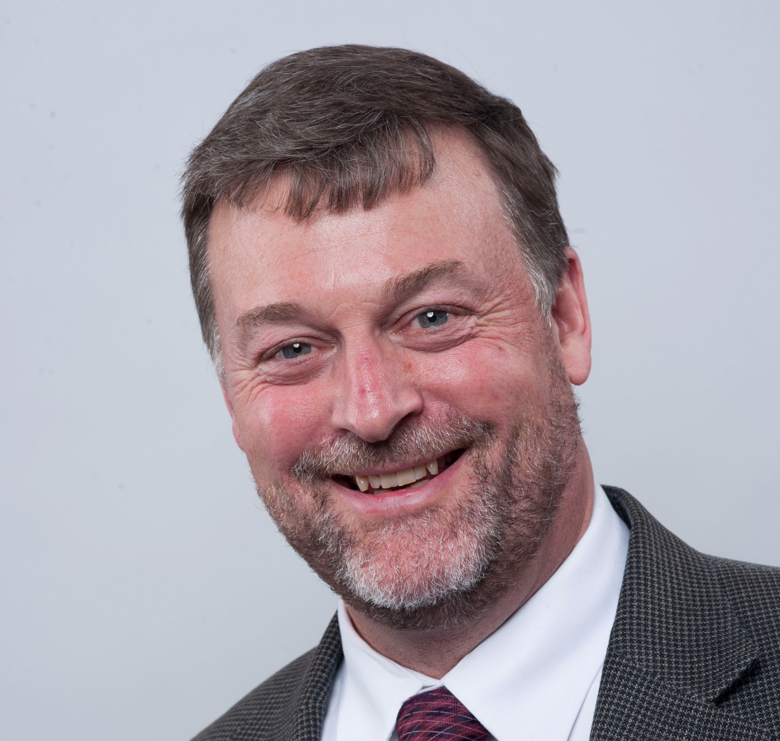Professional headshot of a smiling man with short hair and a beard on a light background